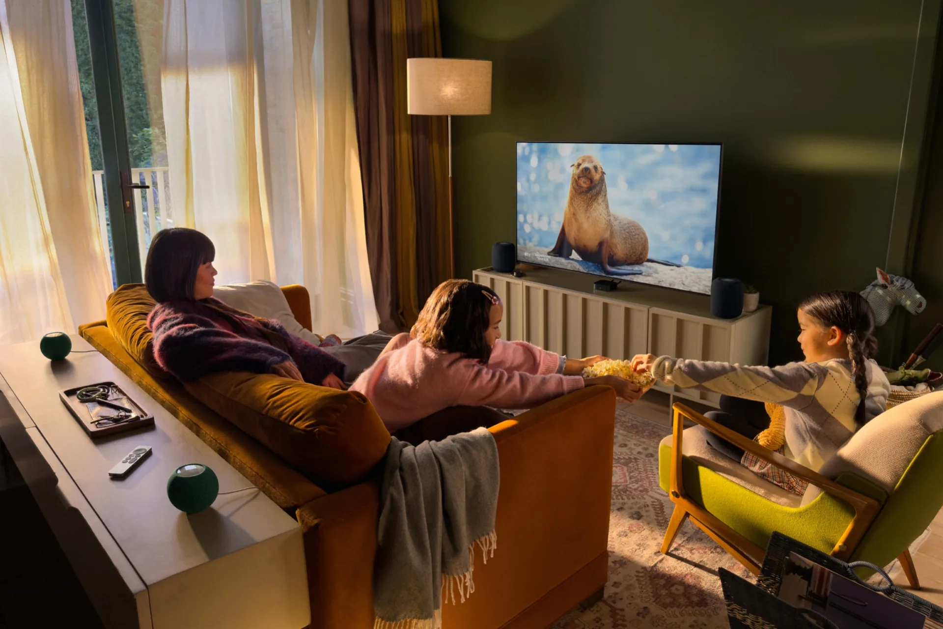 Family watching television in a living room with Apple TV 4K and HomePod mini speakers around the sofa